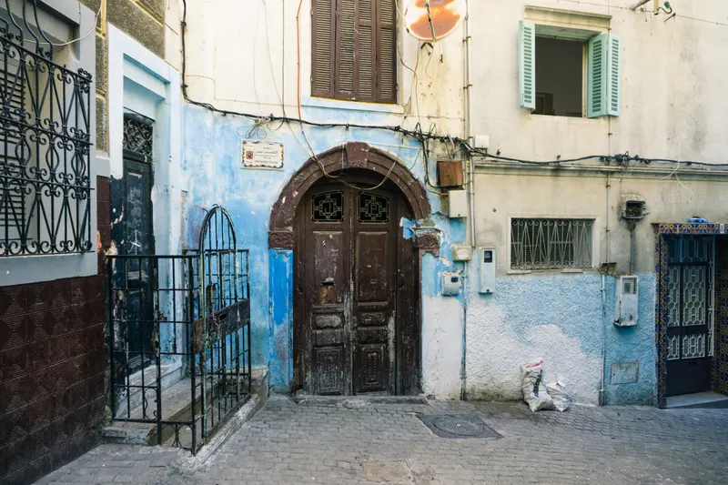 A dark wooden doorway in a weathered white and blue facade, with shutters and overhead wires in Tangier.